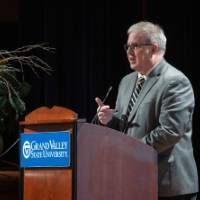 Man speaking to the audience at a GVSU podium, gesturing with one hand